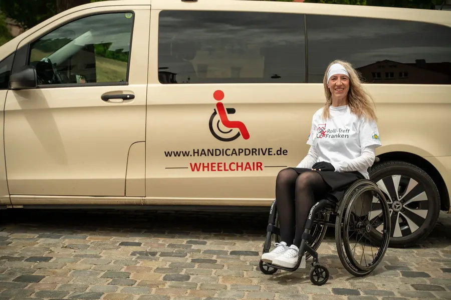 Lächelnde Rollstuhlfahrerin vor einem Handicap-Drive-Fahrzeug