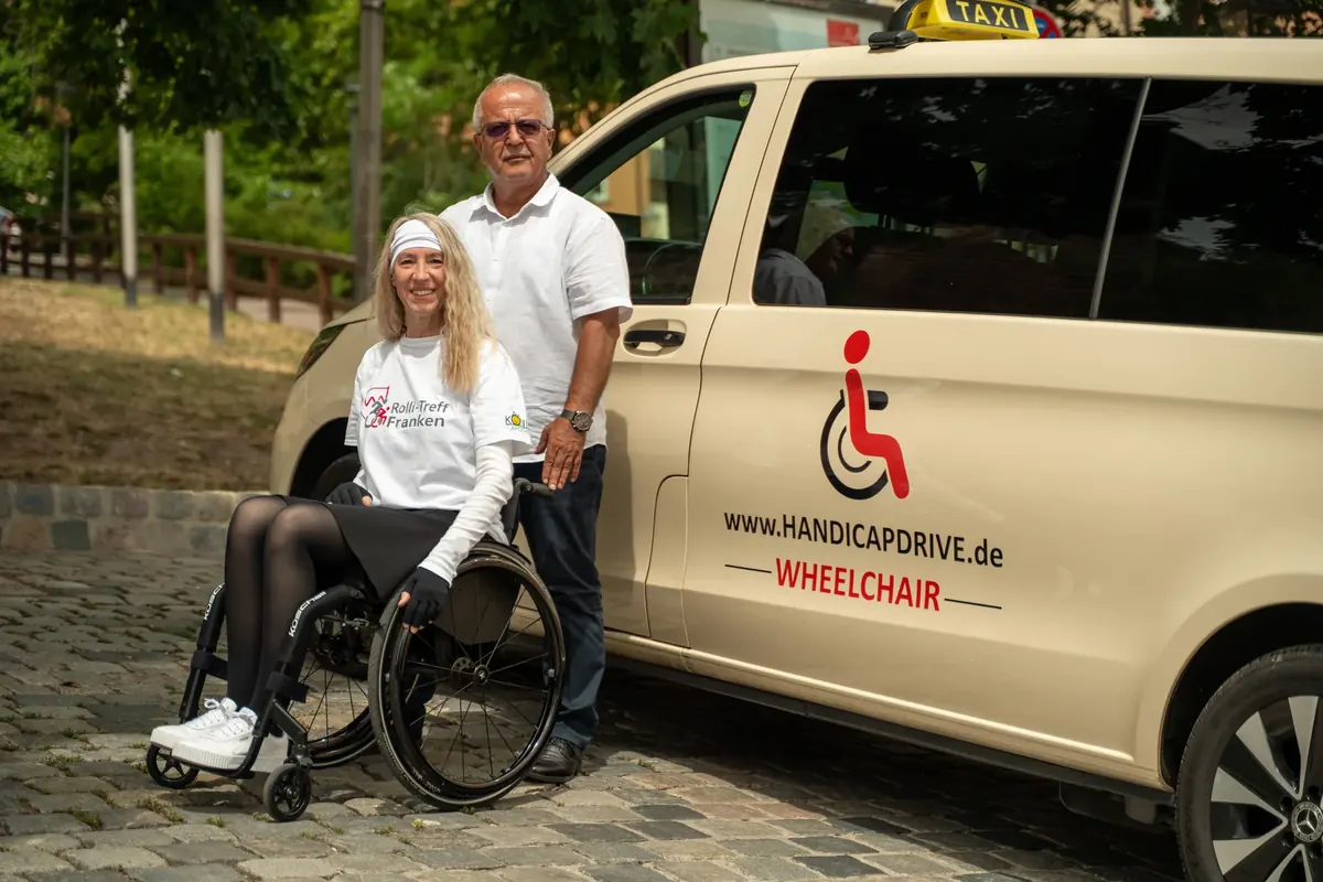 Fahrer und Rollstuhlfahrerin lächeln vor einem Handicap-Drive-Fahrzeug